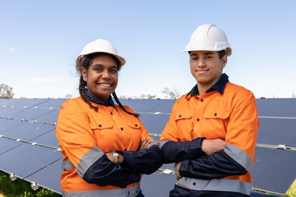 Aboriginal workers in hard hats and bright work gear stand smiling, cross armed, in front of solar panels