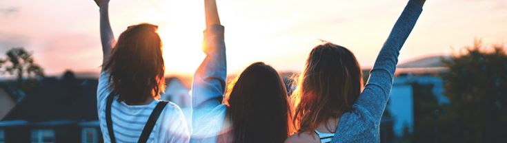 Three young people with their arms raised facing away from the camera looking towards sunrise