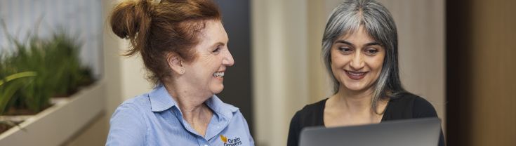 Woman with red hair wearing a blue shirt standing next to a woman with grey hair wearing a black shirt. Both are smiling while working on a grey laptop.