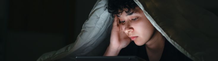 A young person is laying in bed, in the dark watching an iPad. They look sad and tired.