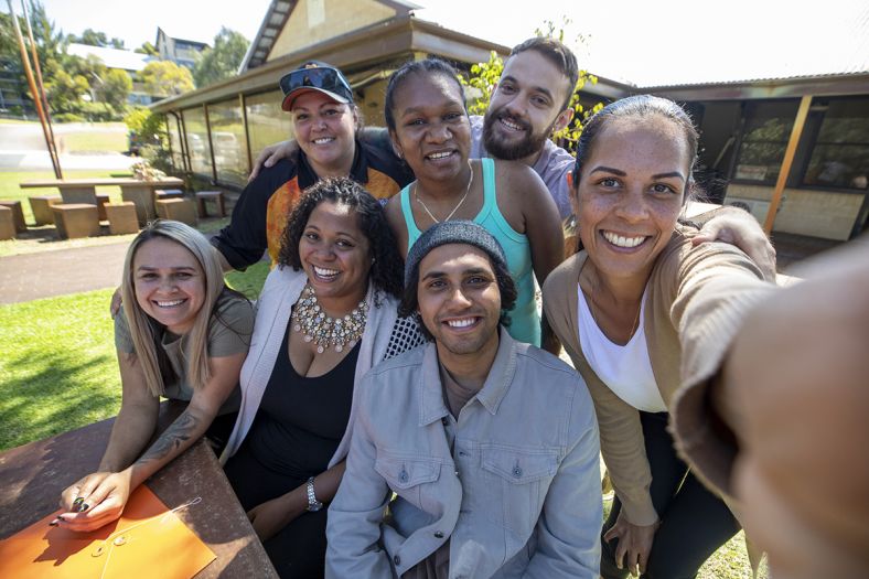 A group of 7 young adults are standing together, posing for a selfie.
