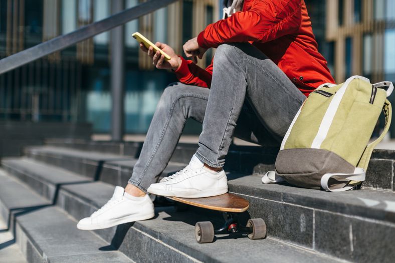 A young person is sitting on outdoor stairs in an urban area holding their phone. Next to them is their backpack and a skateboard.