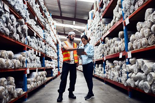 Workers standing between 2 giant warehouse aisles stacked high with rolled up products.