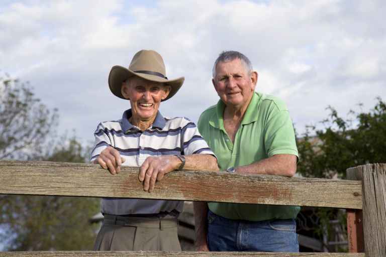 Two men standing on farm land behind a fence, smiling at the camera