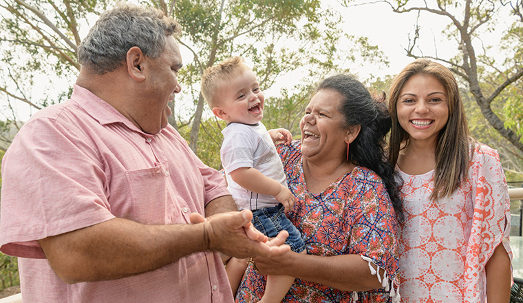 A family of four are standing together in front of the camera smiling. The two elder members are holding a small child who is laughing.
