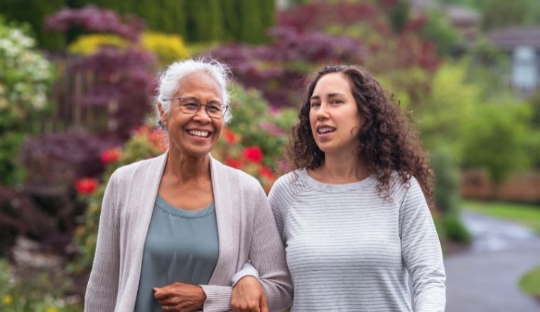 Two women of multicultural background, one is mature aged, the other younger, walking down a path, talking and smiling. 