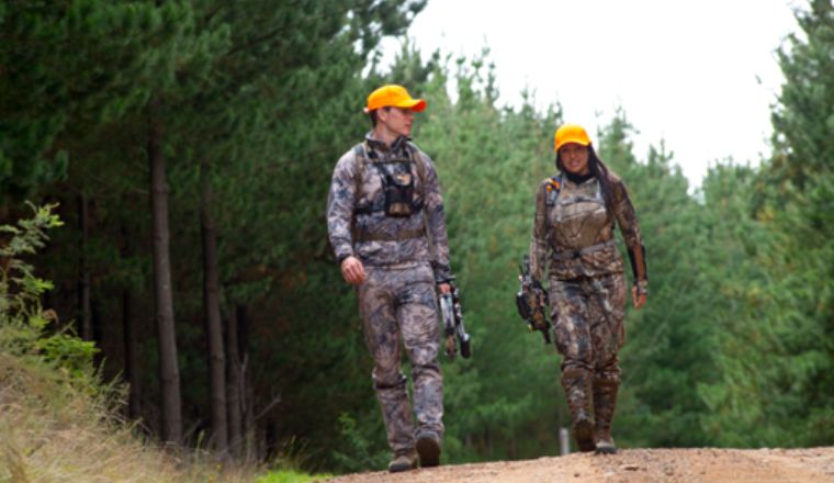 A man and woman wearing camouflage clothes and yellow hats, walking through a forest, carrying guns. 