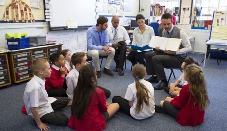 Children sitting on the floor of a classroom, being addressed by a group of adults.