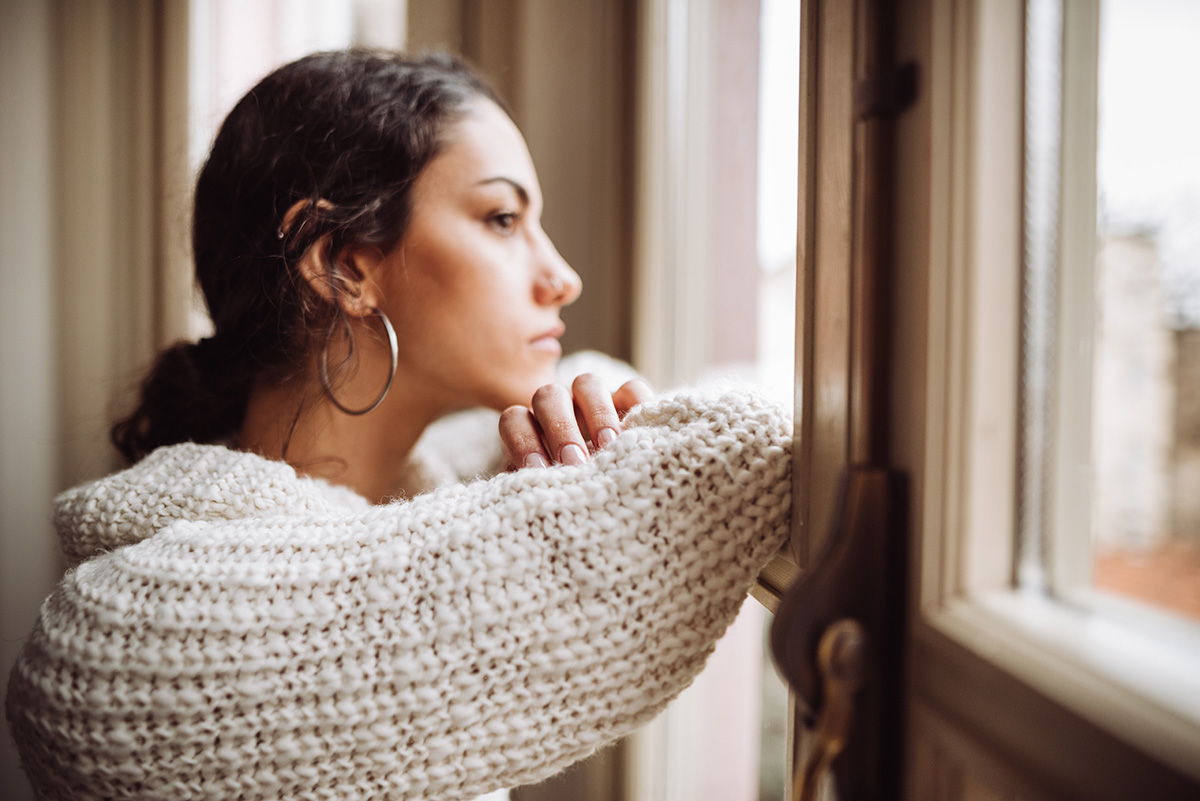 A woman is standing at a window, staring outside looking sad.