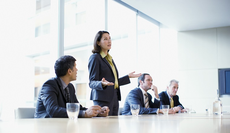 A woman standing up, addressing a meeting of seated colleagues.