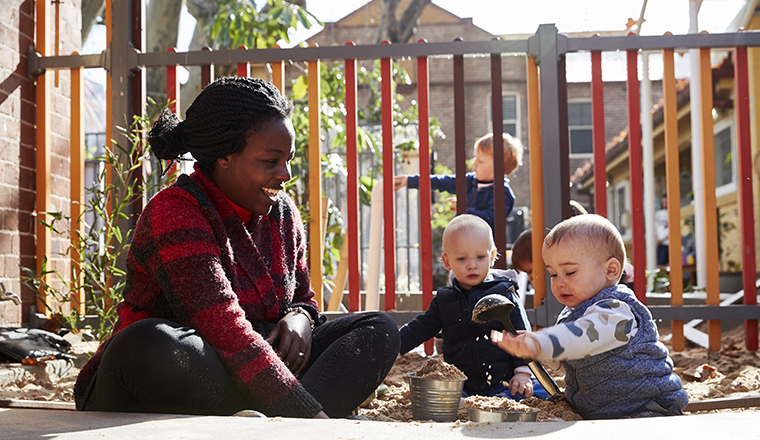Two children play outside in a sandpit accompanied by an early childhood education worker