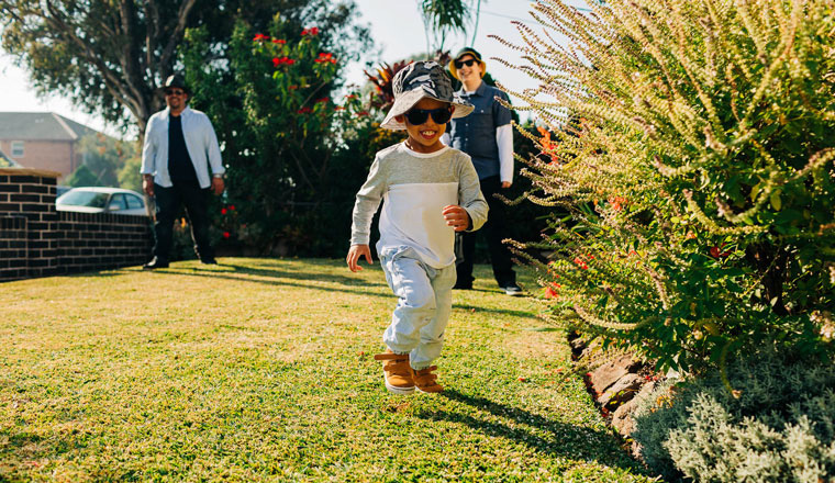 A small child wearing a hat and sunglasses running across a lawn with his parents watching in the background. It is a sunny day.