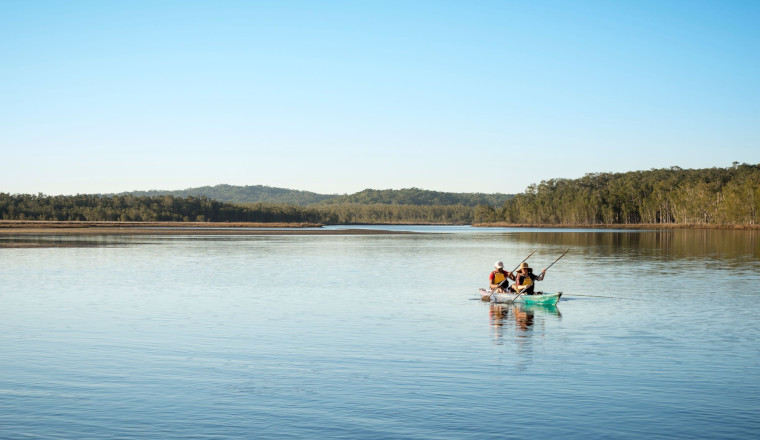 Two people kayaking on a calm lake surrounded by forested hills under a clear blue sky.