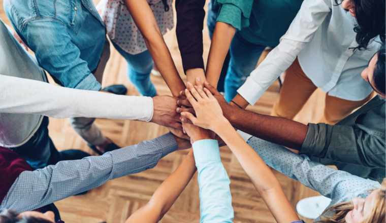 Several people with different skin tones, wearing business casual clothing, stand together in a circle, joining their hands in the middle.