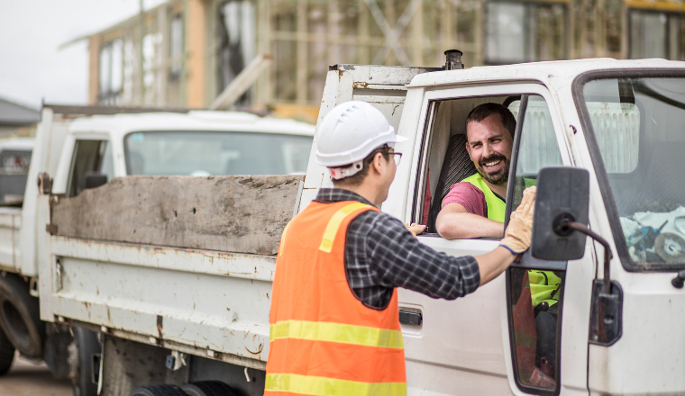A worker in an orange vest and a hard hat talks to another worker who is sitting in a truck. Both are smiling.