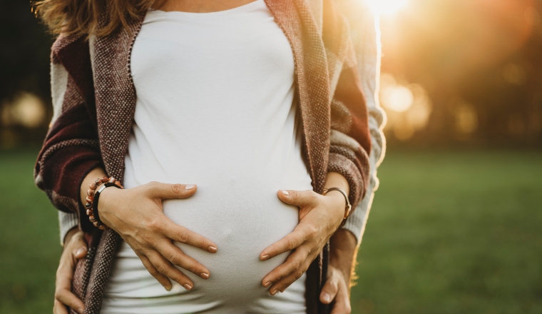 A heavily-pregnant woman clasps her hands around her tummy while her partner embraces her while standing behind.