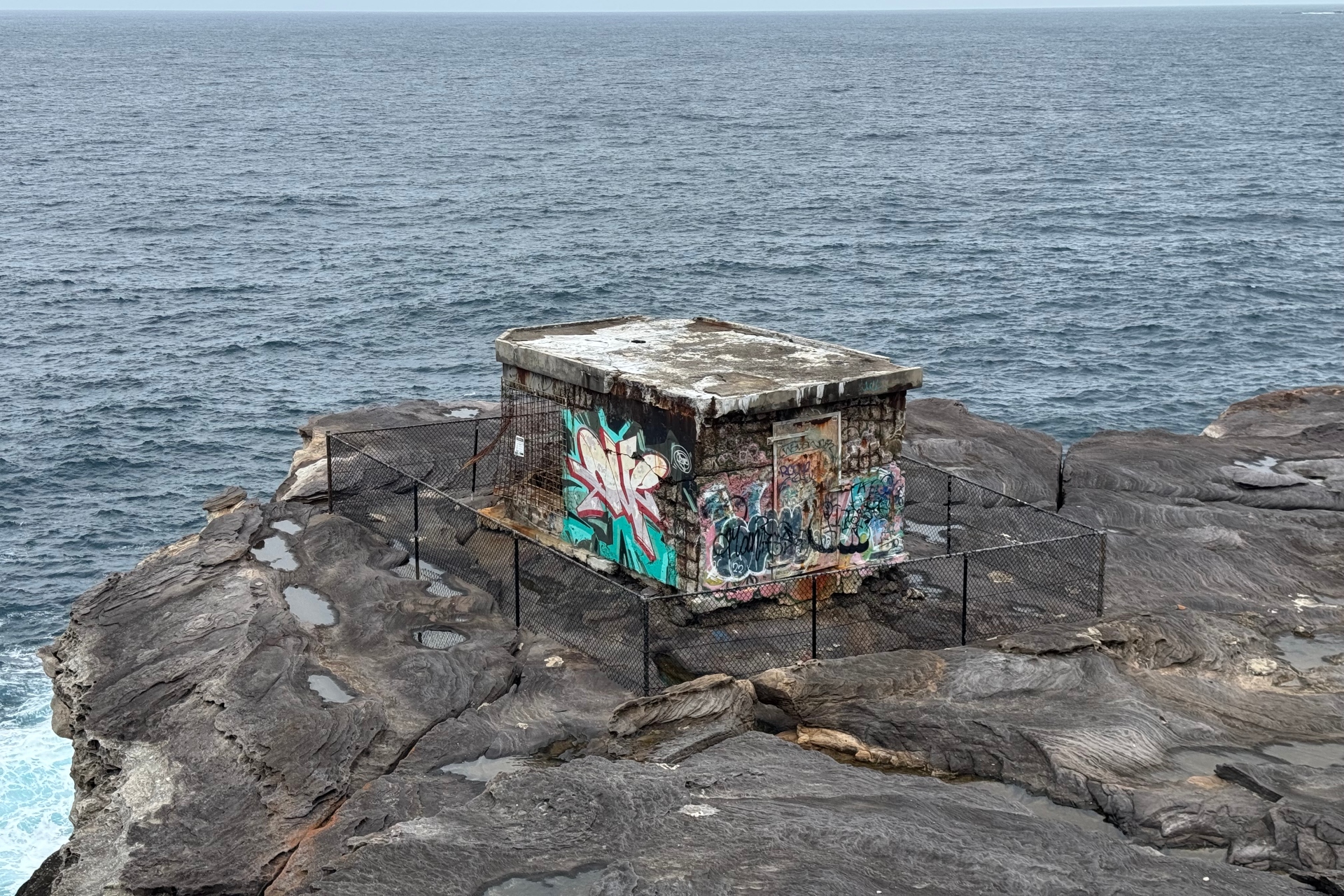 What looks like a graffiti-covered storage shed surrounded by fence, rock and ocean.