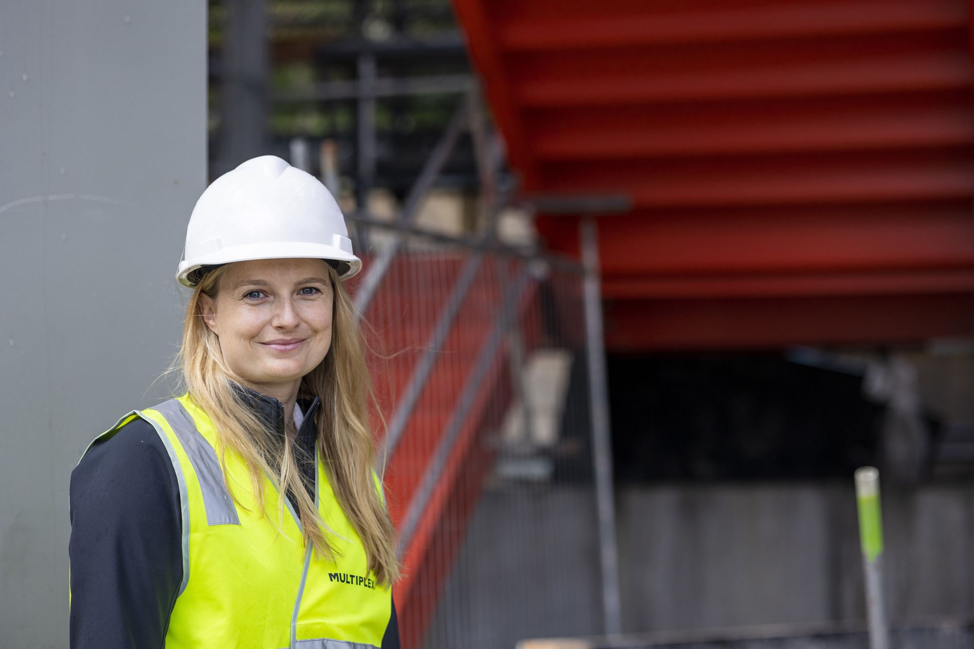 A woman stands on a construction site in hi vis and hard hat. She is smiling at the camera. 