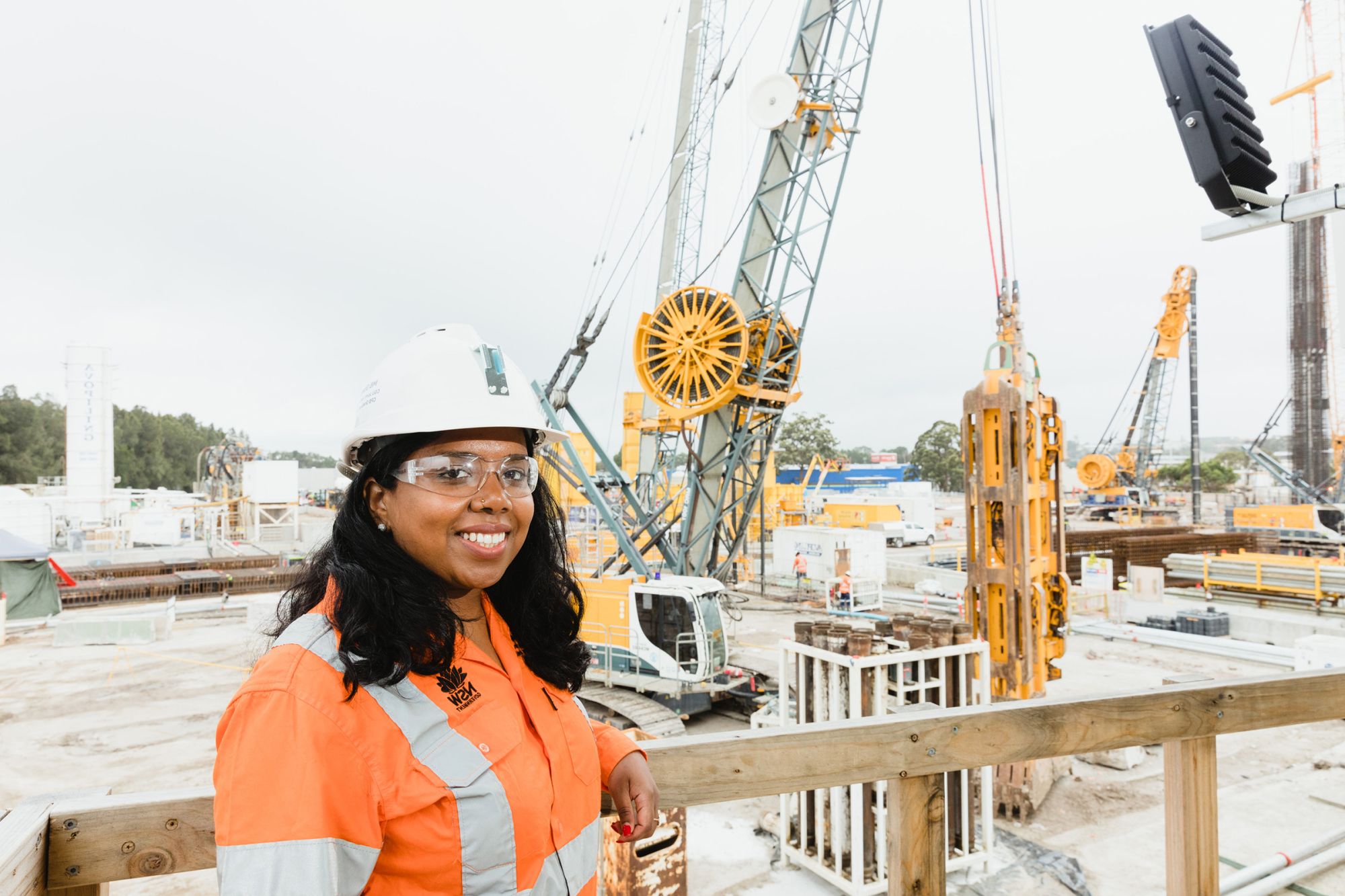 Loshika is standing on a construction site in hi-vis and a hard hat. She is looking at the camera smiling and has machinery behind her.
