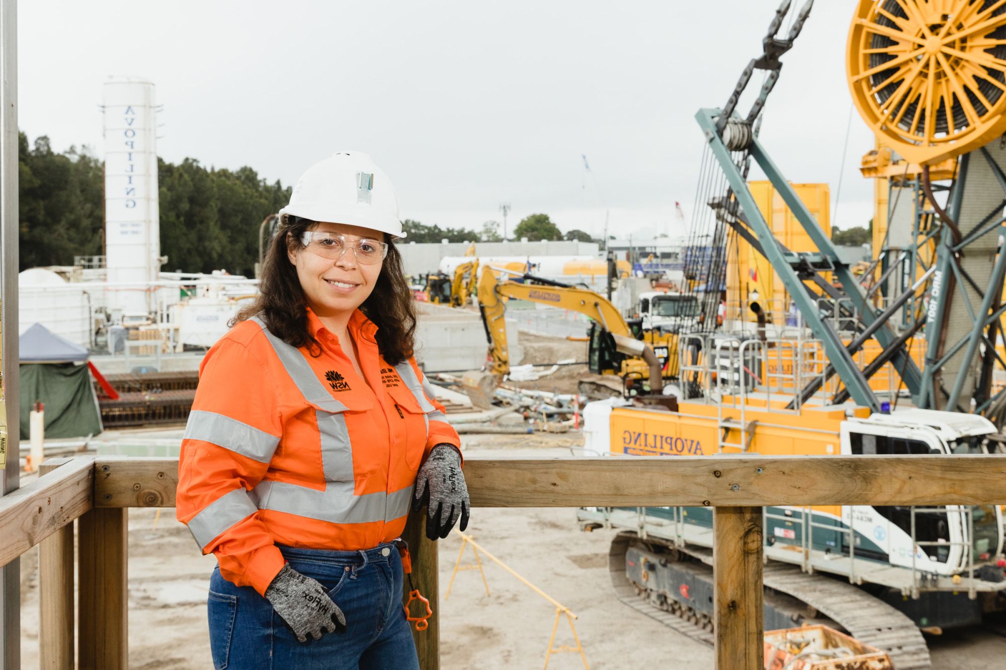 Nagham is standing on site wearing hi vis and a hard hat. She is smiling at the camera and has machinery behind her