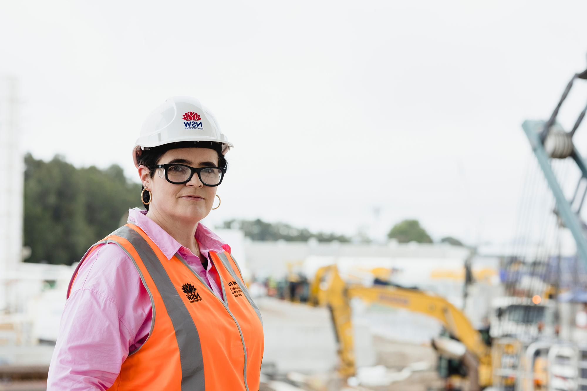 Teresa is standing in front of large machinery wearing a pink t-shirt, hi-vis vest and hard hat.