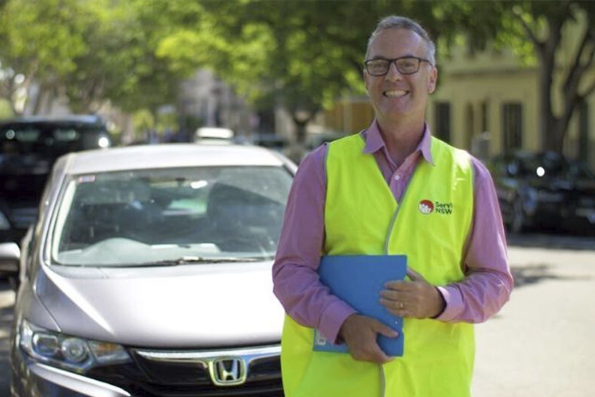 A service NSW driving test supervisor with notepad smiling at the camera in front of a car.