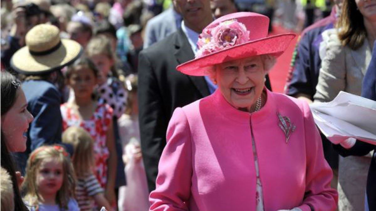 Her Majesty Queen Elizabeth II is walking in a crowd wearing a bright pink jacket and skirt with a matching pink hat.