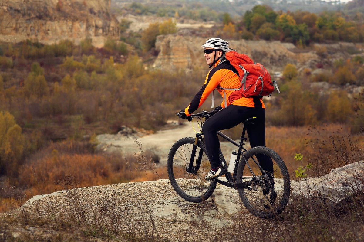 A man in an orange jacket and backpack sit son a bicycle and looks over a brown scrubby landscape