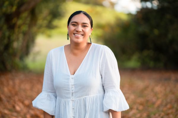 A young Aboriginal woman is standing in a clearing. She is wearing a white blouse  and is smiling candidly at the camera.