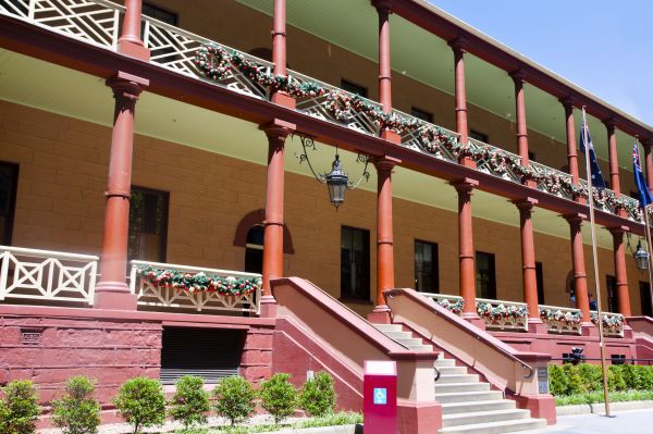 The Macquarie Street frontage of NSW Parliament house in Sydney. The image features an old two story building. The building is a cream colour, but both levels of the building have a balcony and porch that are trimmed with a red/brown paint and white grated metal work. 