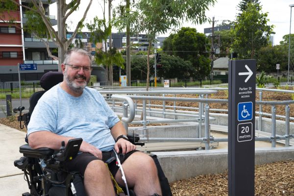 Smiling man with glasses and beard sits in wheelchair next to accessible ramp and sign for Parker St accessible access
