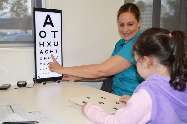 A nurse points to a eye test board with letters to a child sitting with her back to the camera