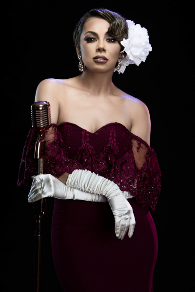 Portrait shot of Prinnie Stevens wearing an embellished maroon strapless dress with a white flower in her hair. She's holding an old-school microphone in her left hand. 
