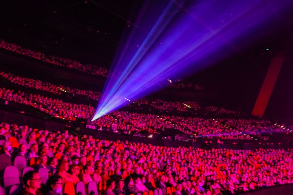 Photo of large crowd of seniors watching a show on stage. A bright spotlight is shining from the middle of the crowd towards the stage. The whole audience is a pink-red hue due to lighting. 