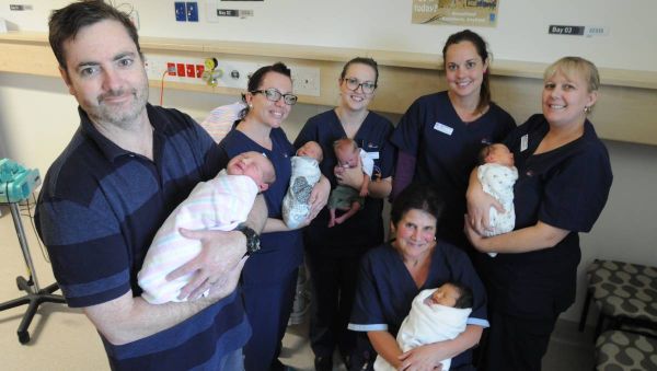 Man and 5 smiling women all holding wrapped newborn babies