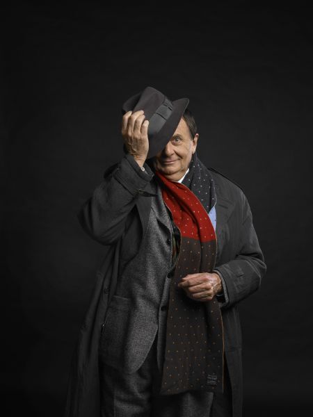 Mr Barry Humphries AO CBE is standing in a dark grey coat against a black background. He is wearing a red scarf and tilting a black hat over one eye smiling.