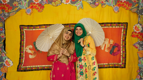 A bright yellow background with two multi lingual story time performers standing in front of it, holding small wooden umbrellas, wearing bright coloured dresses and hijabs.
