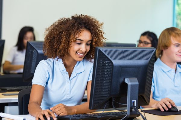 A high school student works on a computer in her school uniform. There are other school students on computers in the background.