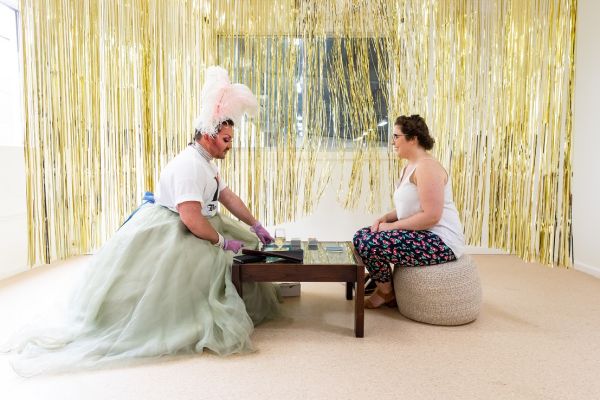 A drag queen performer, Liam Benson and a woman sitting at a table, in front of a gold foil fringe background
