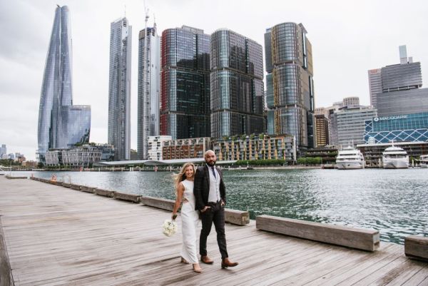 A couple walks along a wooden wharf. Behind them tall modern city buildings reflect the light off the water