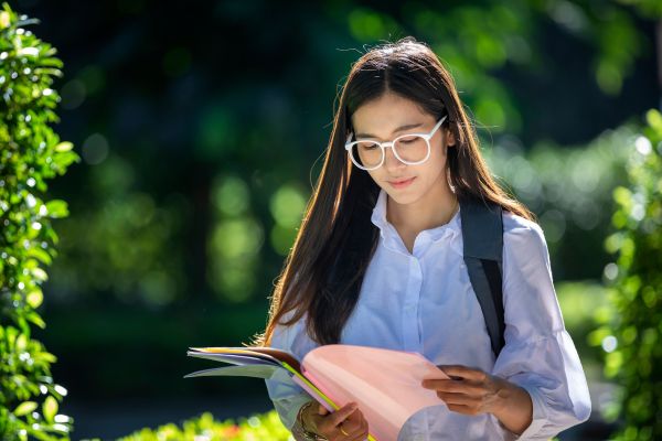 Female senior student reads notes in a leafy park dappled in sunshine. 