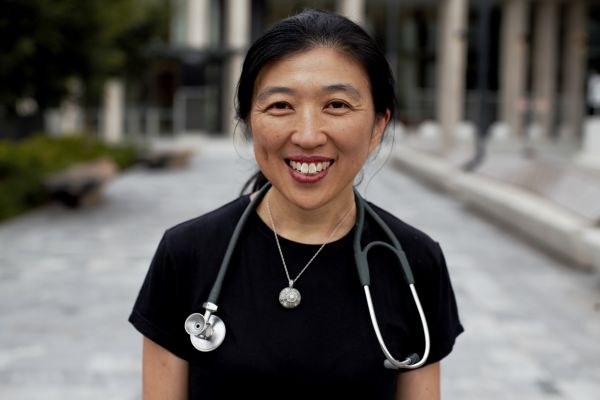 A woman smiles brightly, wearing a black shirt and a stethoscope