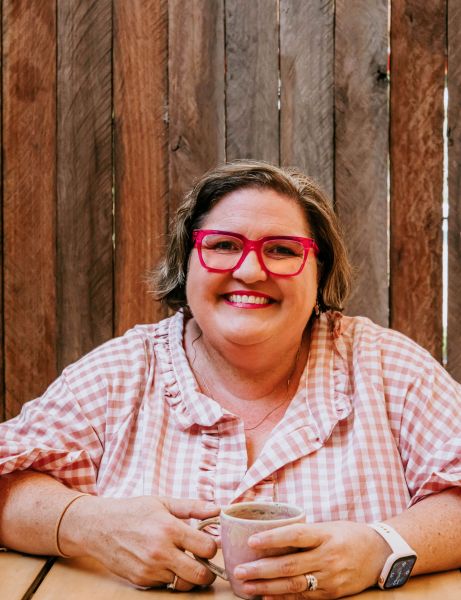 Woman in a pink gingham shirt and pink glasses smiles while holding a mug of coffee