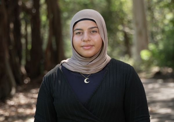 A teenage girl standing outside, wearing a hijab and smiling softly