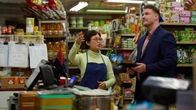 A mature Asian woman talks to a young man in a suit in her grocery store
