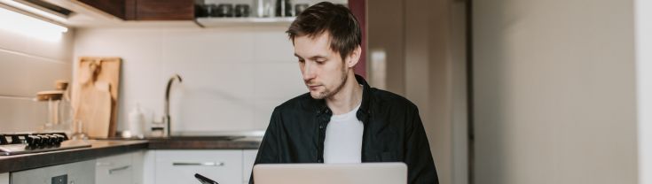 man looks at mobile phone while sitting in front of laptop