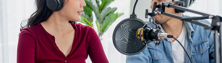 A young woman wearing headphones talking to a young man also wearing headphones behind a microphone which is covered by a pop filter