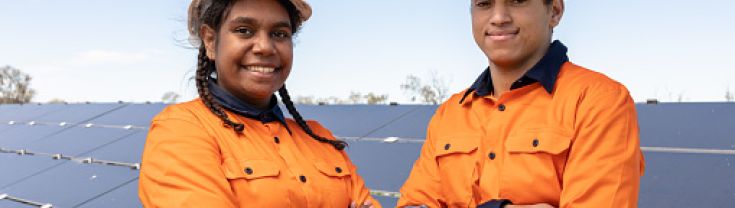 Aboriginal workers in hard hats and bright work gear stand smiling, cross armed, in front of solar panels