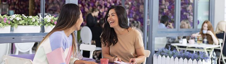 The image shows two women sitting at the Social Hideout Cafe in Parramatta enjoying drinks. The cafe features very bright purple and pink flowers with a neon sign that says 'hello gorgeous' in the background of the image