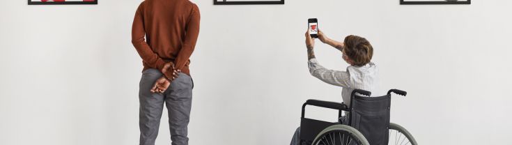 2 men in art gallery looking at paintings one man in wheel chair taking photo